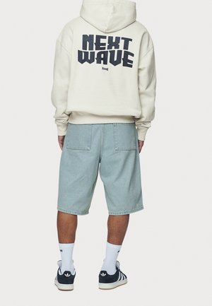 Person trägt einen cremefarbenen Hoodie mit der Aufschrift "NEXT WAVE", hellblaue Jeansshorts, weiße Sportsocken und schwarze Adidas-Sneaker und steht mit dem Rücken zur Kamera.