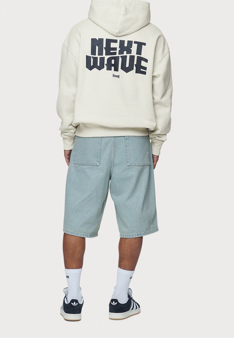 Personne portant un sweat à capuche crème avec le texte "NEXT WAVE", un short en jean bleu clair, des chaussettes blanches montantes et des baskets Adidas noires, debout de dos.