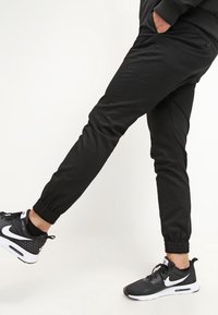 Pantalons de jogging noirs avec des poignets élastiques, tissu lisse, poches latérales, portés avec des baskets Nike noir et blanc présentant un design perforé.