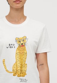 Bílá bavlněná trička s grafikou žlutého leoparda s brýlemi a červenými rty. Text říká "ŠPATNÉ VIBRACE" a "NECHOĎ SE MOU RTĚNKOU."