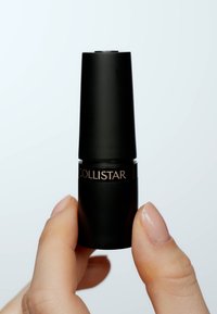 Svart cylindrisk läppstiftstube med en glansig svart finish och en guld "COLLISTAR"-etikett nära basen, hålls mellan två fingrar.