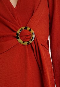 Punainen wrap-top, joka on valmistettu tekstuurikankaasta, ja siinä on kierretty muotoilu sekä suuri keltainen ja musta kilpikonnakuosisormuskoru.