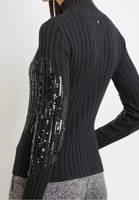 Pull col roulé en maille côtelée noire avec manches longues, orné de paillettes le long des bras. Design texturé et ajusté.