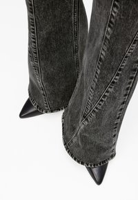 Μαύρο τζιν παντελόνι με φαρδύ τελείωμα και αντίθετη ραφή, αναδεικνύοντας μαύρες μυτερές γόβες στο ύψος του τελειώματος. Ο σχεδιασμός τονίζει την πλατιά γραμμή και την υφή του υφάσματος.