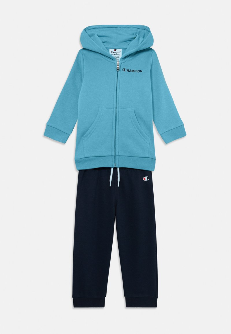 Sudadera azul con cremallera y bolsillos, acompañada de pantalones de chándal azul marino. Ambos hechos de una tela suave, con el logo de "Champion" en el pecho y la pierna del pantalón.