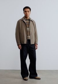 Άνδρας που φοράει μπεζ σακάκι πάνω από καφέ ριγέ πουκάμισο, φαρδιά σκούρα τζιν και καφέ παπούτσια, στέκεται μπροστά σε ένα απλό φωτεινό γκρι φόντο.