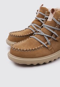 Scarpe in suede marrone con punta rotonda, cuciture testurizzate e lacci grigi. La suola è di un beige chiaro con un design ondulato.