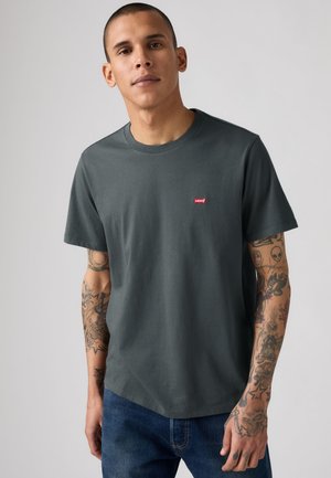 Jeune homme portant un t-shirt gris foncé Levi's et un jean bleu, montrant des tatouages détaillés couvrant les deux bras.