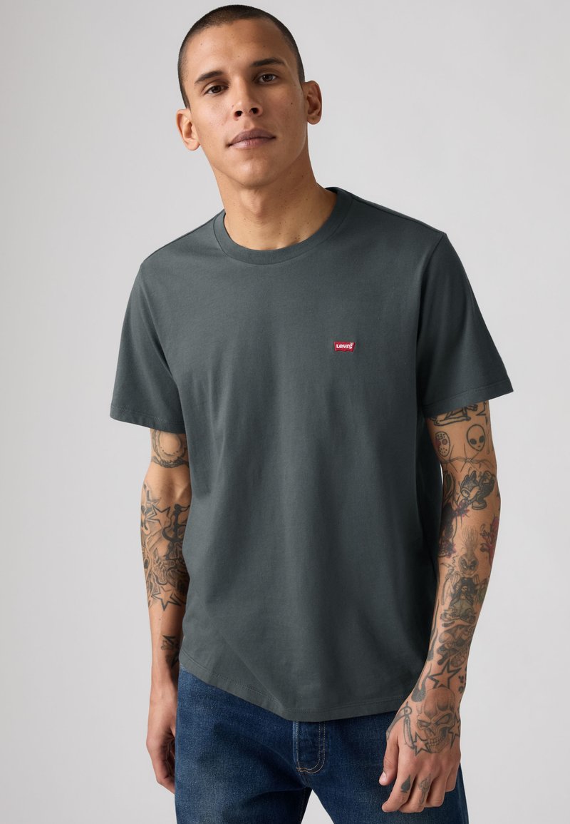 Jeune homme portant un t-shirt gris foncé Levi's et un jean bleu, montrant des tatouages détaillés couvrant les deux bras.