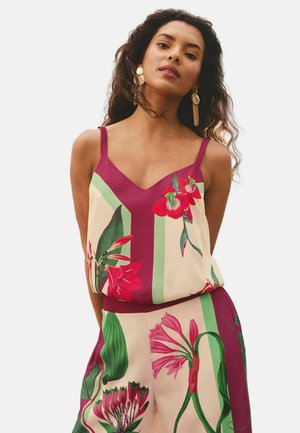 Nő, aki ujjatlan virágmintás felsőt és hozzáillő nadrágot visel piros, rózsaszín és zöld növénymintákkal, nagy arany fülbevalóval kiegészítve.