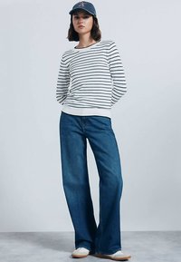 Felpa a righe bianca e blu navy, vestibilità comoda, abbinata a jeans blu scuro a gamba larga. Il modello indossa un berretto blu navy e scarpe chiare.