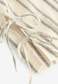 Vêtement en tissu naturel avec base crème et motif à rayures noires ; comporte des liens décoratifs et des détails de tissage texturé.