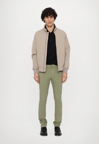 Beige bomberjacka med ribbad mudd, svart pikétröja under, ljusgröna slim-fit byxor och svarta skor. Framsidan, neutral bakgrund.