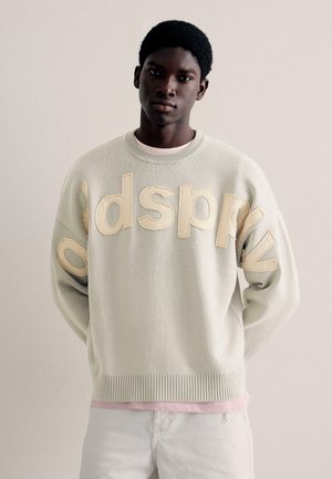 Jonge man met kort haar die een beige trui draagt met grote gestikte letters over de borst, staand tegen een effen lichte achtergrond.