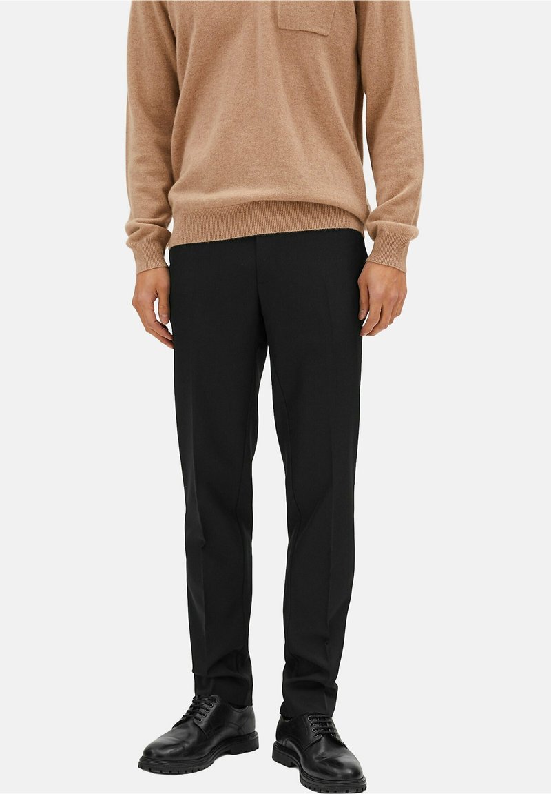 Homme portant un pull beige, un pantalon noir sur mesure et des chaussures en cuir noires, debout devant un fond uni.