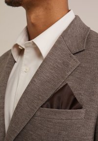 Brauner Textur-Blazer mit einem satinbraunen Einstecktuch, kombiniert mit einem weißen Hemd mit Kragen. Auffällige Details sind die scharfen Revers und der Knopfverschluss.