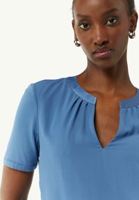 Jasnoniebieska bluzka z krótkim rękawem, z dekoltem w kształcie litery V i miękką fakturą. Posiada marszczenia przy dekolcie dla dodatkowego wykończenia.