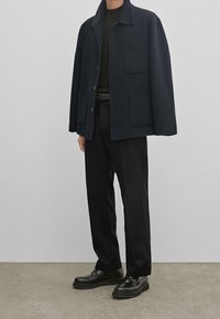 Giacca di lana blu navy con due tasche frontali, indossata sopra una maglietta scura, abbinata a pantaloni neri su misura e scarpe nere lucide.