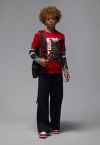 T-shirt graphique rouge avec un design représentant un trophée, manches longues camouflées, pantalon de survêtement noir et chaussures de sport rouges avec des accents blancs.