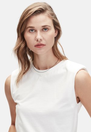Witte mouwloze top met een ronde halslijn, gemaakt van zachte stof. Model met schouderlang haar draagt een eenvoudige zwarte ketting.