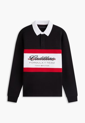 Jonge man die een zwart, rood en wit poloshirt met lange mouwen draagt met de tekst "Cadillac Formula 1 Team", witte broek, zwarte schoenen en een witte pet.