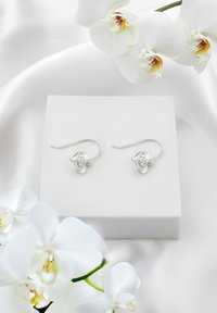 Söljearmband med blomsterdesign, med en liten central ädelsten, som visas på en vit låda mot silkesliknande tyg och vita orkidéer.