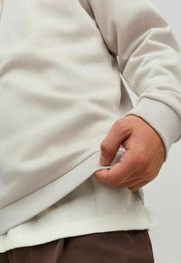 Sudadera de punto gris claro con puños y dobladillo acanalados. Una mano sostiene el borde inferior, revelando un contraste de texturas con una capa oculta debajo.