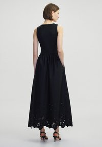 Schwarzes ärmelloses Kleid mit tailliertem Oberteil und ausgestellt Rock. Verfügt über bestickte florale Ausschnitte am Saum. Getragen mit Riemchen-Highheels.