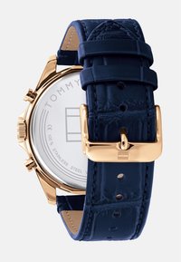 Orologio con cinturino in pelle blu e cassa rotonda in acciaio inossidabile. Presenta dettagli dorati, una superficie liscia e un quadrante bianco semplice con logo.