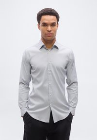 Άνδρας με κοντά σγουρά μαλλιά που φοράει ανοιχτό γκρι πουκάμισο με κουμπιά και μαύρο παντελόνι, στέκεται με τα χέρια στις τσέπες μπροστά σε απλό λευκό φόντο.