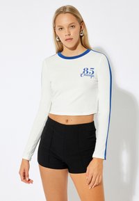Witte cropped lange mouwen shirt met blauwe accenten en "85 Chicago" gedrukt in blauw. Gecombineerd met hoge zwarte shorts.