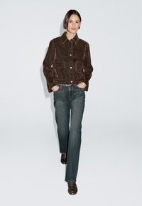 Vrouw die loopt, gekleed in een donkergouden corduroy jas, rechte donkerblauwe jeans en donkergouden instaplaarzen, met haar handen in haar zakken tegen een effen lichte achtergrond.