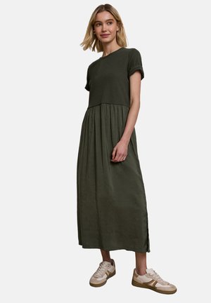 Jeune femme portant une robe maxi à manches courtes vert foncé et des baskets blanches, debout devant un fond clair uni.
