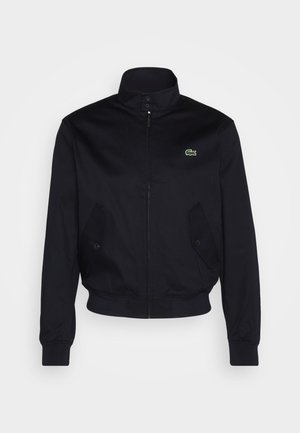 Marinblaue Jacke mit hohem Kragen, Reißverschluss vorne und zwei Seitentaschen; mit grünem Lacoste-Logo auf der Brust; aus glattem Stoff gefertigt.