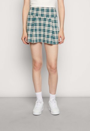 Vrouw draagt een blauw-witte geruite geplooide minirok, witte sokken en witte sneakers, staand tegen een effen lichte muur.
