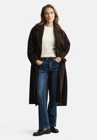 Langer, dunkelbrauner Wollmantel mit breiten Revers, getragen über einem hellen Pullover, kombiniert mit blauen Schlagjeans und schwarzen Slippern.