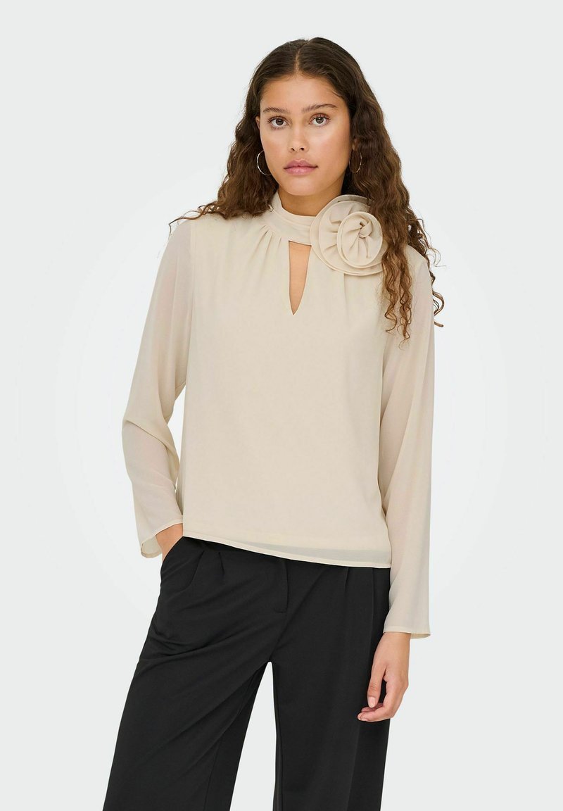 Femme aux longs cheveux bouclés portant un chemisier beige avec une encolure goutte d'eau et une grande décoration florale, associée à un pantalon noir.