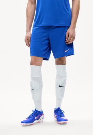 Dolna część ciała osoby noszącej niebieskie szorty sportowe, białe nagolenniki i niebieskie korki piłkarskie z różowymi i białymi akcentami.