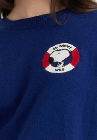 Blauwe trui met een ronde geborduurde patch met Snoopy en tekst op een witte en rode achtergrond. Zacht breiwerk, ronde hals.
