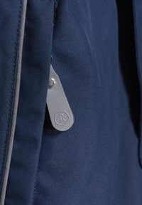 Navy tyg ryggsäck i närbild, med en silver dragkedja med logotyp, strukturerad yta och kontrasterande grå sömmar längs sömmarna.