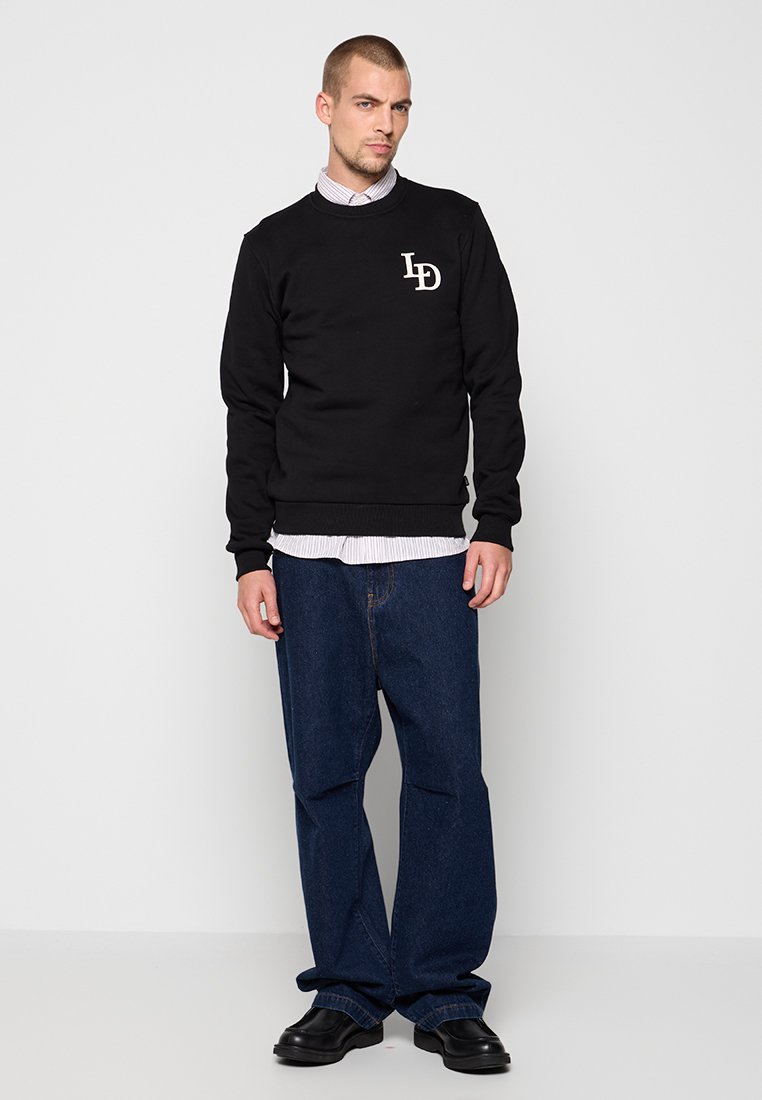 Mann, der einen schwarzen Sweatshirt mit "LD"-Logo, ein gestreiftes Hemd, weite blaue Jeans und schwarze Schuhe trägt, steht vor einem schlichten weißen Hintergrund.