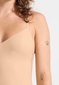 Hudfärgad badkläder med justerbara remmar, som har en slät tygkvalitet och en vågformad kant. En liten tatuering syns på överarmen.
