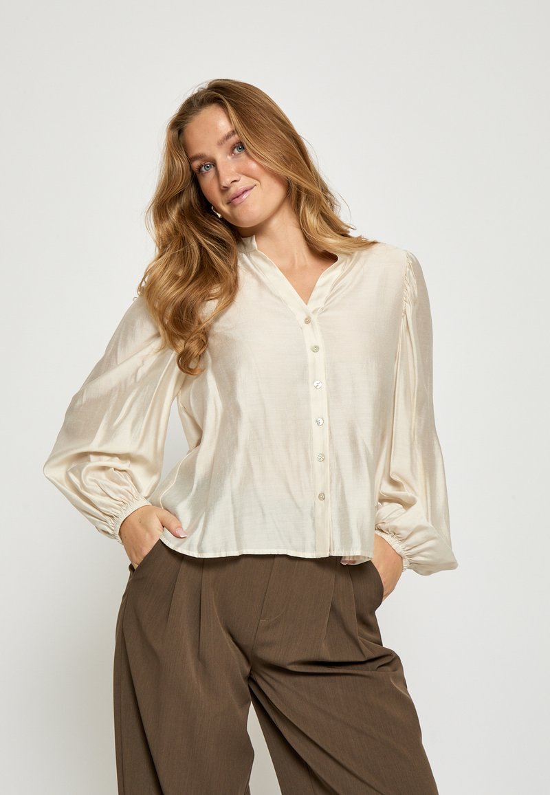 Krēmkrāsas, zīda blūze ar V-veida kaklu, pogu priekšpusi un plandošām piedurknēm. Iezīmēts ar elastīgiem apročām un brīvu piegriezumu.