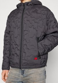 Gepolsterte Kapuzenjacke in Dunkelgrau mit einem gesteppten Muster, frontalem Reißverschluss und seitlichen Taschen. Kleine rote Logoakzente am Saum.