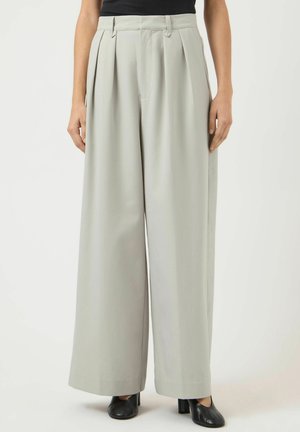 Kvinne iført høytlivs, vidbente lysegrå bukser med folder, kombinert med svarte sko med hæl og en svart topp.