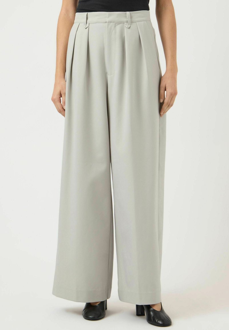 Naine, kes kannab kõrge vöökohaga, laiade säärtega helehallid püksid voldidega, millele on lisatud mustad kontsakingad ja must top.