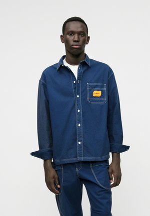 Mørkeblå denimskjorte med lange ærmer, knappet foran, brystlomme med en gul etiket og kontrastsyede detaljer.