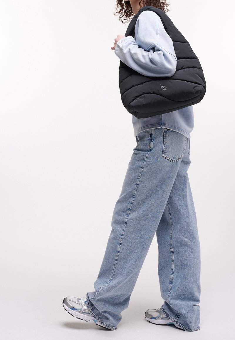 Personne portant un sweat-shirt bleu clair et un jean bleu ample, portant un sac à épaule noir matelassé, marchant en baskets blanches.