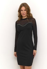 Vestido negro con mangas largas transparentes, escote redondeado y un cuerpo texturizado que presenta una línea ondulada de tachuelas decorativas. Acabado de tela suave.