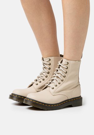Stivali da combattimento in pelle beige con caviglia alta, punta rotonda, suola in gomma nera e cuciture gialle. Allacciati con occhielli in metallo.